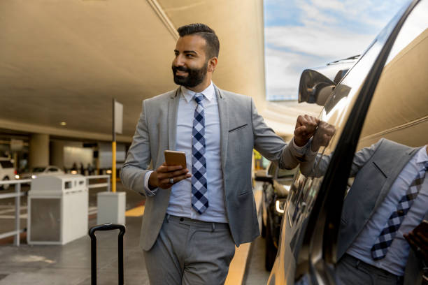 A man in a suit stands next to a car, holding his phone, ready for airport transportation.